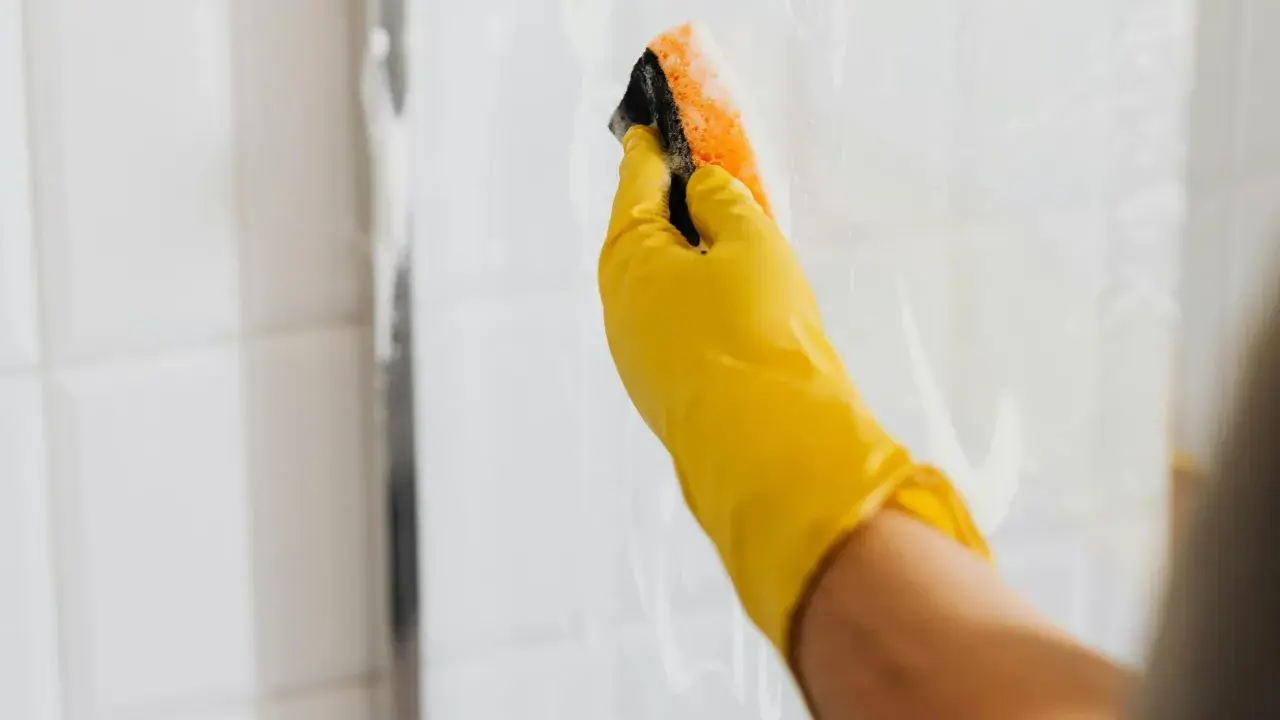 Ręka w żółtej rękawiczce czyści kabinę prysznicową gąbką. Skuteczne czyszczenie kabiny prysznicowej octem i sodą to domowy sposób na lśniące płytki.
