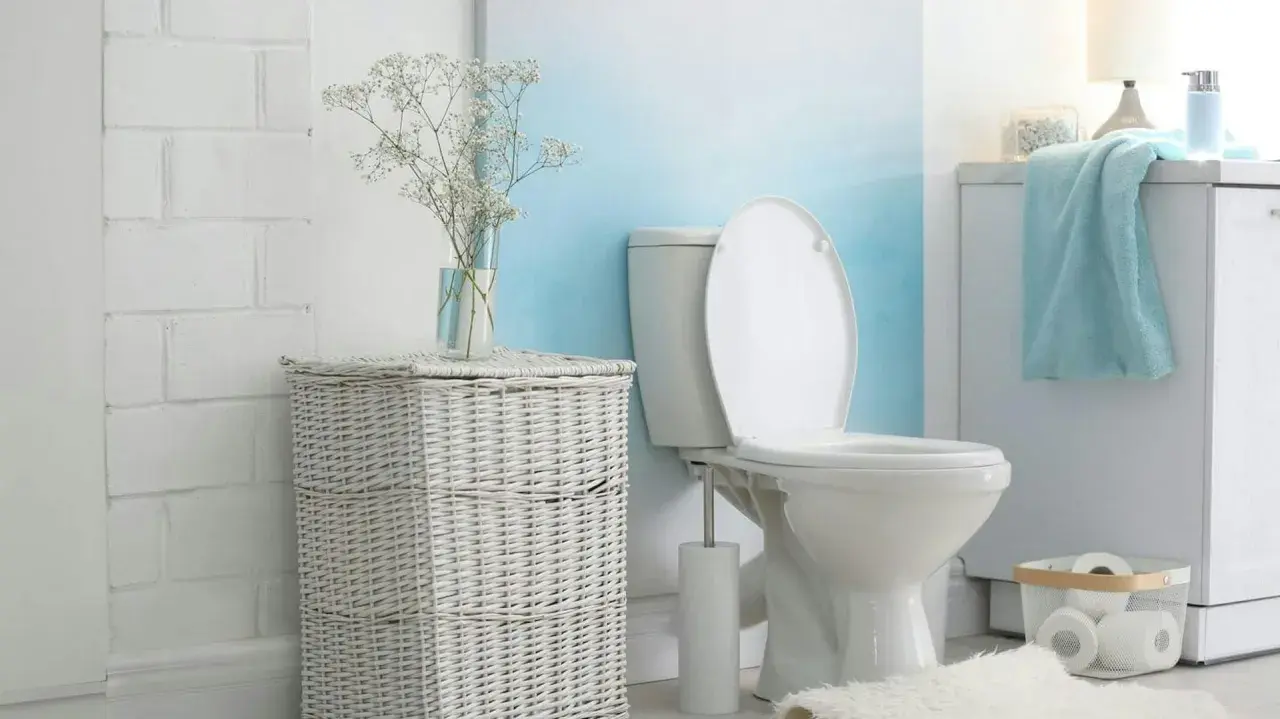 Biała, ceramiczna spłuczka WC starego typu w łazience z wiklinowym koszem i bukietem gipsówki.