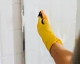 Ręka w żółtej rękawiczce czyści kabinę prysznicową gąbką. Skuteczne czyszczenie kabiny prysznicowej octem i sodą to domowy sposób na lśniące płytki.