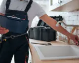 Instrukcja: jak wymienić uszczelkę w kranie. Hydraulik w ogrodniczkach trzyma teczkę, obok zlew i narzędzia.