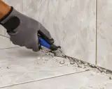 Ręka w rękawicy usuwa starą fugę z narożnika płytek.