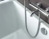 Bateria termostatyczna z chromowanymi elementami, z której strony ciepła woda, nad białą wanną.