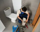Hydraulik naprawia rozwalony kibel. Widać narzędzia i części.