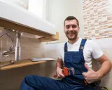 Uśmiechnięty hydraulik w niebieskim ogrodniczkach trzyma klucz nastawny, gotowy do czyszczenia odpływu pod umywalką.