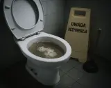 Zatkany kibel z papierem i wodą. Obok stojak "Uwaga dezynfekcja" i przepychacz, który pomoże jak odetkać kibel.
