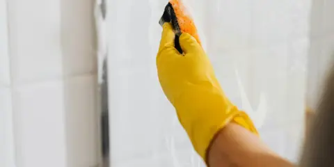 Ręka w żółtej rękawiczce czyści kabinę prysznicową gąbką. Skuteczne czyszczenie kabiny prysznicowej octem i sodą to domowy sposób na lśniące płytki.