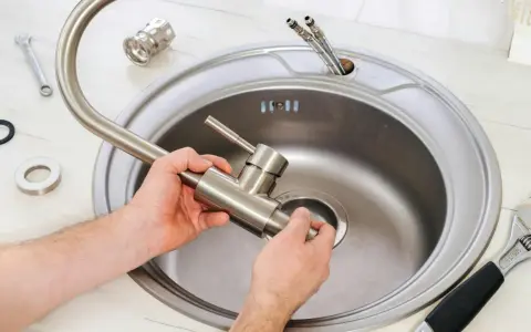 Montaż baterii zlewozmywakowej stojącej. Dłonie trzymają nową baterię, przygotowując ją do instalacji w zlewie. Obok leżą klucze i uszczelki.