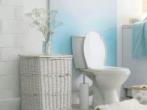 Biała, ceramiczna spłuczka WC starego typu w łazience z wiklinowym koszem i bukietem gipsówki.