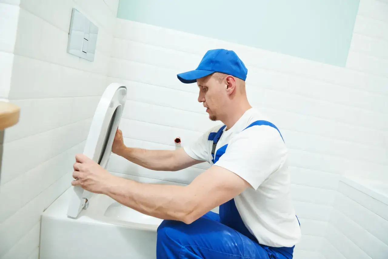 Instalator w niebieskim kombinezonie pokazuje, jak odkręcić deskę sedesową. Białe płytki i jasne wnętrze łazienki.